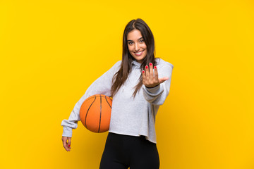 Young woman playing basketball over isolated yellow background inviting to come with hand. Happy that you came