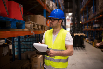 Warehouse worker in reflective uniform with hardhat checking products on shelf in large distribution storage area.