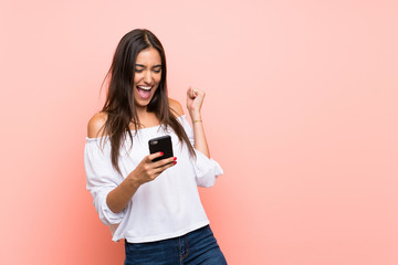 Young woman over isolated pink background with phone in victory position