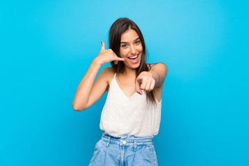 Young woman over isolated blue background making phone gesture and pointing front