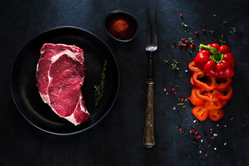 Raw Steak Ribeye on a plate, fork for meat, herbs and spices on a dark concrete background