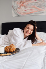 Obraz na płótnie Canvas Young cheerful woman in white bathrobe laughing lying in bed with delicious pastry on breakfast near at modern hotel