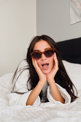 Young attractive woman with dark hair in white bathrobe and sunglasses leaning on hands dreamily looking in camera on big bed in modern cozy hotel