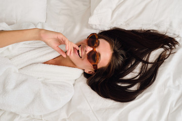 Young gorgeous smiling woman with dark hair in white bathrobe and sunglasses dreamily bite finger...