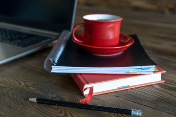 Pencil in front of the red cup of fresh morning coffee on the notobook on wooden table in the office. Coffee break. Top-side view. 