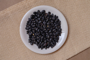 Black beans seeds in a white ceramic cup on a cream colored tablecloth on a brown paper background.
