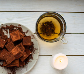 handmade souffle cubes covered with cocoa powder lie on a plate on a light wooden table, hot tea in a glass mug and next to it three white burning candles, pieces of chocolate