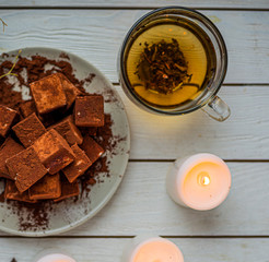 handmade souffle cubes covered with cocoa powder lie on a plate on a light wooden table, hot tea in a glass mug and next to it three white burning candles, pieces of chocolate