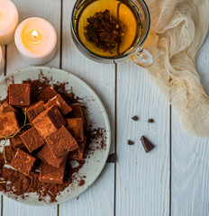 handmade souffle cubes covered with cocoa powder lie on a plate on a light wooden table, hot tea in a glass mug and next to it three white burning candles, pieces of chocolate