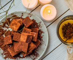 handmade souffle cubes covered with cocoa powder lie on a plate on a light wooden table, hot tea in a glass mug and next to it three white burning candles, pieces of chocolate