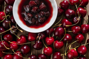 Cherry jam in a small bowl close-up on a wooden background, spread around a lot of ripe juicy cherries