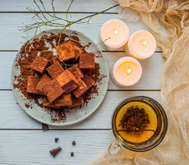 handmade souffle cubes covered with cocoa powder lie on a plate on a light wooden table, hot tea in a glass mug and next to it three white burning candles, pieces of chocolate and a sprig.