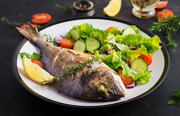 Baked fish dorado with lemon and fresh salad in white plate on dark rustic background. Healthy dinner with fish concept. Dieting and clean eating