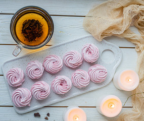 pink handmade zephyr, marshmallow on a light wooden table, hot tea in a glass mug and next to it three white burning candles and pieces of chocolate. Top view