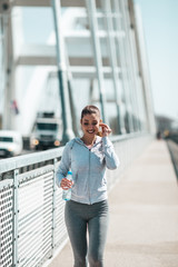 Fototapeta premium Beautiful young and fit woman in good shape running and jogging alone on city bridge street. She wears sporty earbuds and listens to music.
