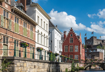 Fototapeta na wymiar Gent, Flanders, Belgium - June 21, 2019: Narrow Lieve River leads along old bourgeois mansions to Gravensteen Castle under blue sky with white clouds. Bluish water. Flowers on quays.