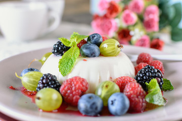 Delicious cream Panna Cotta dessert decorated with various berries mint leaves are at a plate on a table, next to tea cup and flowers
