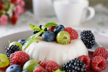 Delicious cream Panna Cotta dessert decorated with various berries mint leaves are at a plate on a table, next to tea cup and flowers