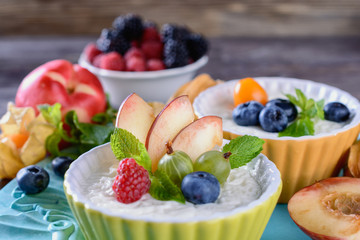 Delicious cream Panna Cotta dessert decorated with various berries, fruits and mint leaves in ceramic form on a wooden table next to ingredients, closeup