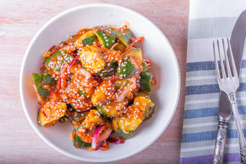 Oi Muchim Korean spicy cucumber salad with sesame seeds in a bowl and fork with knife on a wooden table, top view.