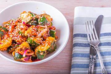 Oi Muchim Korean spicy cucumber salad with sesame seeds in a bowl and fork with knife on a wooden table, top view.