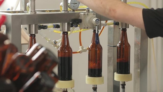 At a brewery, a machine fills a bottles of beer. A machine operator controls the process. Close-up.