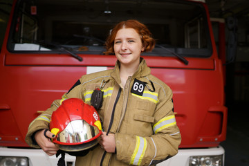 Photo of smiling woman firefighter with helmet in hands against backdrop of fire truck