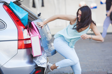 Attractive young girl trying to put a lot of retails into a car. Woman putting shopping bags into a...