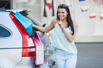 Attractive young girl trying to put a lot of retails into a car. Woman putting shopping bags into a...