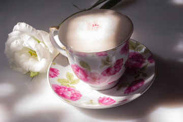A cup of coffee cappuccino with floral print on a white table, where the shadows and rays of the morning sun and a white eustoma flower, the concept of waking up a morning cup of coffee.