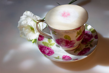 A cup of coffee cappuccino with floral print on a white table, where the shadows and rays of the morning sun and a white eustoma flower, the concept of waking up a morning cup of coffee.