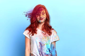 Young cheerful red-haired girl under explosion of colored powder at Holi colors. close up portrait, isolated blue background, studio shot.