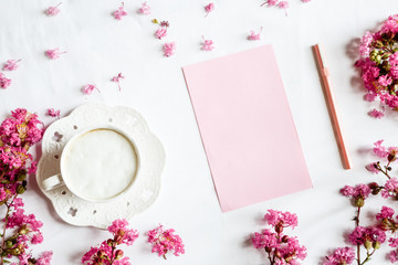 Morning coffee mug for breakfast, empty notebook and pink flowers on white wooden table, top view, flat lay style. Woman working desk. Mockup, overhead