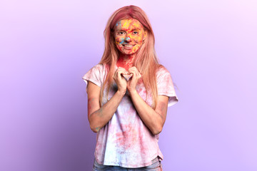 Beautiful cheerful fair-haired woman with colored face and clothes makes heart symbol by hands. I love Holi. emotion and feeling