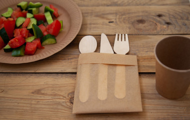 paper cardboard dishes with vegetable salad tomatoes cucumbers with wooden devices fork horse knife on a wooden table