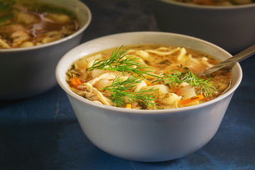 Nourishing Chicken Noodle Soup White Bowl Closeup. Full Dish of Starter Meal. Healthy Family Recipe. Poultry Bouillon with Chopped Veggies and Herbs. Warm Homemade Lunch on Kitchen Table