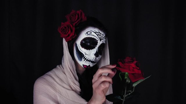 Halloween mask CALAVERA CATRINA. Girl with a red rose. Mexican day of the dead. Portrait of a young woman with scary multi-colored makeup for Halloween on a dark background. 4K