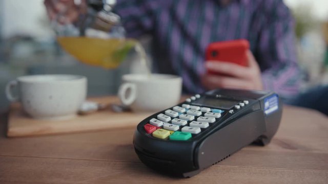 Young man sitting in the cafe pouring tea into two cups. Close-up of customer paid for order with paypass technology on cash mashine lying on table.