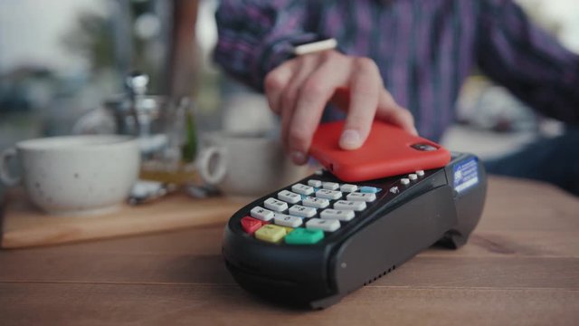 Close-up of cash machine and young man using smartphone for quick paypass payment sitting by table indoors. Young man customer paying for order eating on break in the cafe.