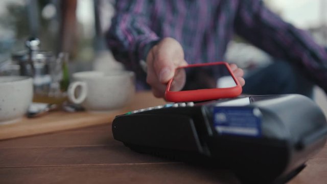 Close-up male customer paying for order with nfc technology on smartphone while sitting in the cafe on coffee break.