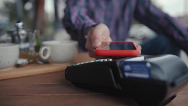 Contactless NFC technology. Close-up young man client sitting by table using mobile phone paypass to pay for order putting a device on cash machine.