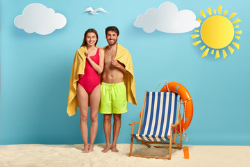 Travel and summer concept. Glad couple shelter under soft beach towel, dressed in swimsuit, hug together with love, empty deck chair with lifebuoy near, hide from sun, share intimate moment.