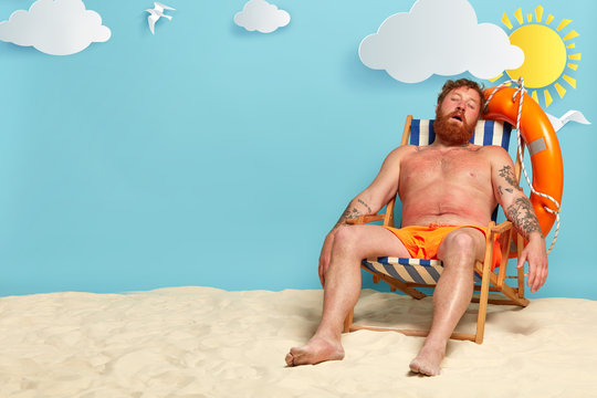 Relaxed bearded man sleeps on his deck chair, poses on beach, has red sunburned skin, sunbathes and tans, has summer vacation and holiday travel, beautiful sea view in background. People and rest