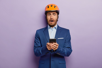 Frightened builder man wears formal suit and hardhat, holds mobile phone, reacts on getting message, has widely opened eyes, stands against purple background. Engineer with modern cellular in hands