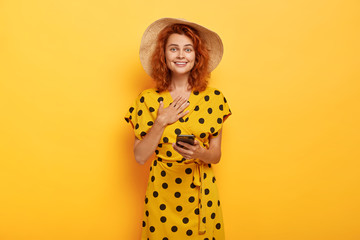 Satisfied impressed red head woman touches chest, wears summer hat and yellow polka dot dress,...