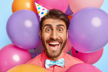 Lucky cheerful man with happy look, makes photo against party attributes, wears cone paper hat,...