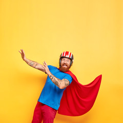 Funny cheerful male superhero wears helmet and red cloak, going to fall down, keeps arms outstretched, gazes at camera, isolated on yellow background with empty space above. Heroes still exists © WHstudio Leushin N