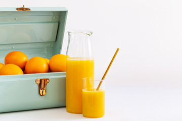 Orange juice in a glass and a jug by a blue metal small suitcase full of oranges over a white background.