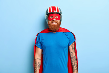 Glad bearded man with funny outlook, comes on costume party, being superhero character, wears helmet, mask and red cloak, has fun with friends, poses against blue background. Save world concept