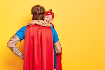 Lovely glad kid embraces tightly father who stands back to camera, wears red cloak, plays together...
